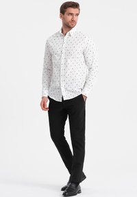 Camisa blanca de botones con patrón abstracto negro, de mangas largas y con cuello puntiagudo. Combinada con pantalones negros ajustados y zapatos negros.