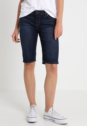 Shorts Bermuda en denim bleu foncé avec une coupe ajustée, taille mi-haute et ourlets repliés, associés à des baskets blanches et un t-shirt blanc.