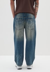 Jeans de mezclilla azul de corte holgado con parches desgastados, dos bolsillos traseros y diseños de estilo graffiti en blanco. Llevados con una camisa marrón y zapatillas blancas.