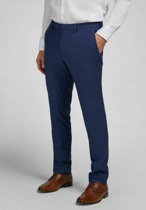 Mann trägt marineblaue, schmal geschnittene Stoffhose, weißes Hemd und braune Lederschuhe, steht mit einer Hand in der Hosentasche.