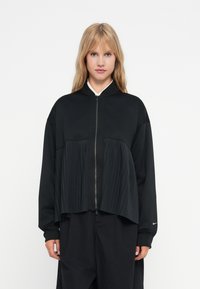 Casaco preto com fecho éclair, apresentando um design plissado na frente, gola alta e ombros caídos. Fabricado em tecido suave e liso.