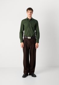Cămașă verde cu mânecă lungă, cu un design ajustat, asortată cu pantaloni maro plisati și o curea neagră cu o cataramă logo din argint.