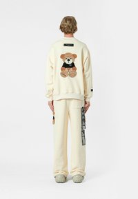 Sudadera oversize de color crema con un aplique texturizado de oso de peluche en la espalda, combinada con pantalones a juego y zapatos grises gruesos.