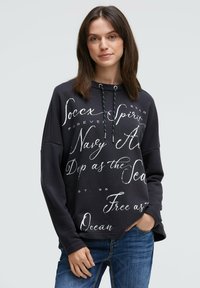 Schwarzer Kapuzenpullover mit weißen Texten in verschiedenen Schriftarten, langen Ärmeln und lockerer Passform. Kombiniert mit blauen Jeans.