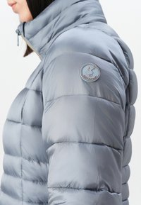 Vrouw in een zilvergrijze pufferjack met hoge kraag en een ronde "Save The Duck" logo patch op de bovenste linkermouw.