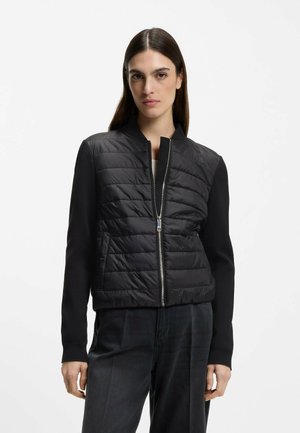 Femme portant une veste matelassée noire à fermeture éclair avec des manches en tricot et un pantalon taille haute foncé, debout devant un fond blanc uni.