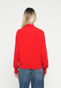 Kobieta z długimi blond włosami, ubrana w czerwoną bluzę i niebieskie jeansy, stojąca plecami w stosunku do jednolitego białego tła.