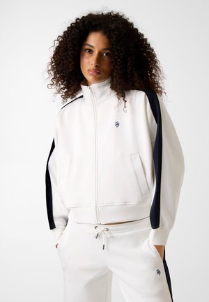 Persoon met krullend haar, gekleed in een wit trainingspak met zwarte zijstrepen, met handen in de zakken, staand tegen een effen achtergrond.