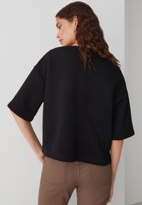 Felpa nera cortissima con maniche ampie, realizzata in tessuto morbido, presenta un design pulito e dettagli di cucitura minimi, indossata su pantaloni marroni.