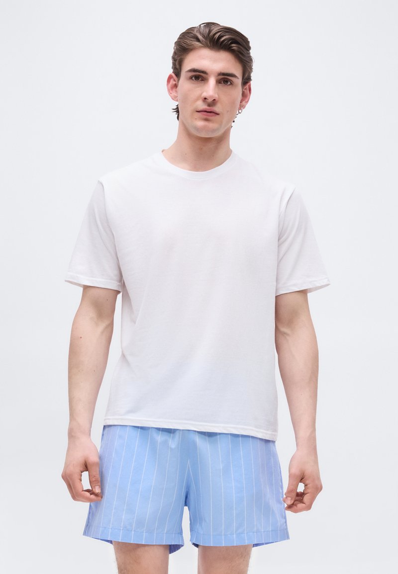 Νεαρός άνδρας με κοντά καστανά μαλλιά, φορώντας απλό λευκό t-shirt και ανοιχτό μπλε σορτς με λεπτές ρίγες, στέκεται μπροστά σε απλό φόντο.