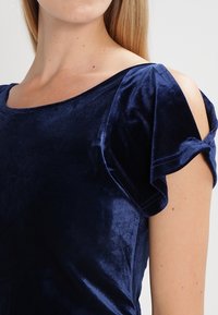 Granatowa aksamitna bluzka z krótkimi, falbaniastymi rękawami i dekoltem w serek. Materiał ma miękkie, teksturowane wykończenie.