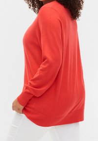Pull en maille corail à manches longues avec poignets et ourlet côtelés, doté d'une coupe décontractée et d'une texture douce, porté avec un pantalon blanc.