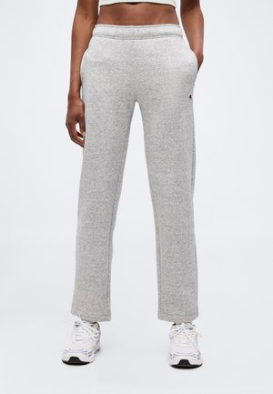 Vrouw die lichtgrijze joggingbroek met rechte pijpen en zijzakken draagt en witte sneakers, getoond van taille tot voeten op een effen achtergrond.