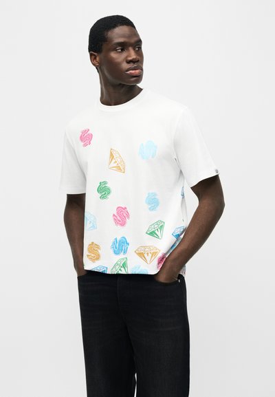 Άνδρας που φοράει λευκό μπλουζάκι με πολύχρωμα γραφικά δολαρίων και διαμαντιών, στέκεται με τα χέρια στις τσέπες, κοιτάζοντας προς τα πλάγια.