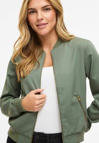 Veste bomber vert olive en tissu lisse, dotée d'une fermeture éclair à l'avant et de poches latérales, avec poignets et ourlet côtelés.