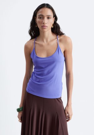 Femme aux longs cheveux foncés portant un haut violet sans manches et une jupe marron, debout devant un fond clair uni.
