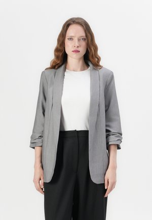 Šedý blazer s ohrnutými rukávy, zapínání na jeden knoflík a měkkou texturou, nošený přes bílou košili a černé kalhoty. Jednoduchý, přizpůsobený design.