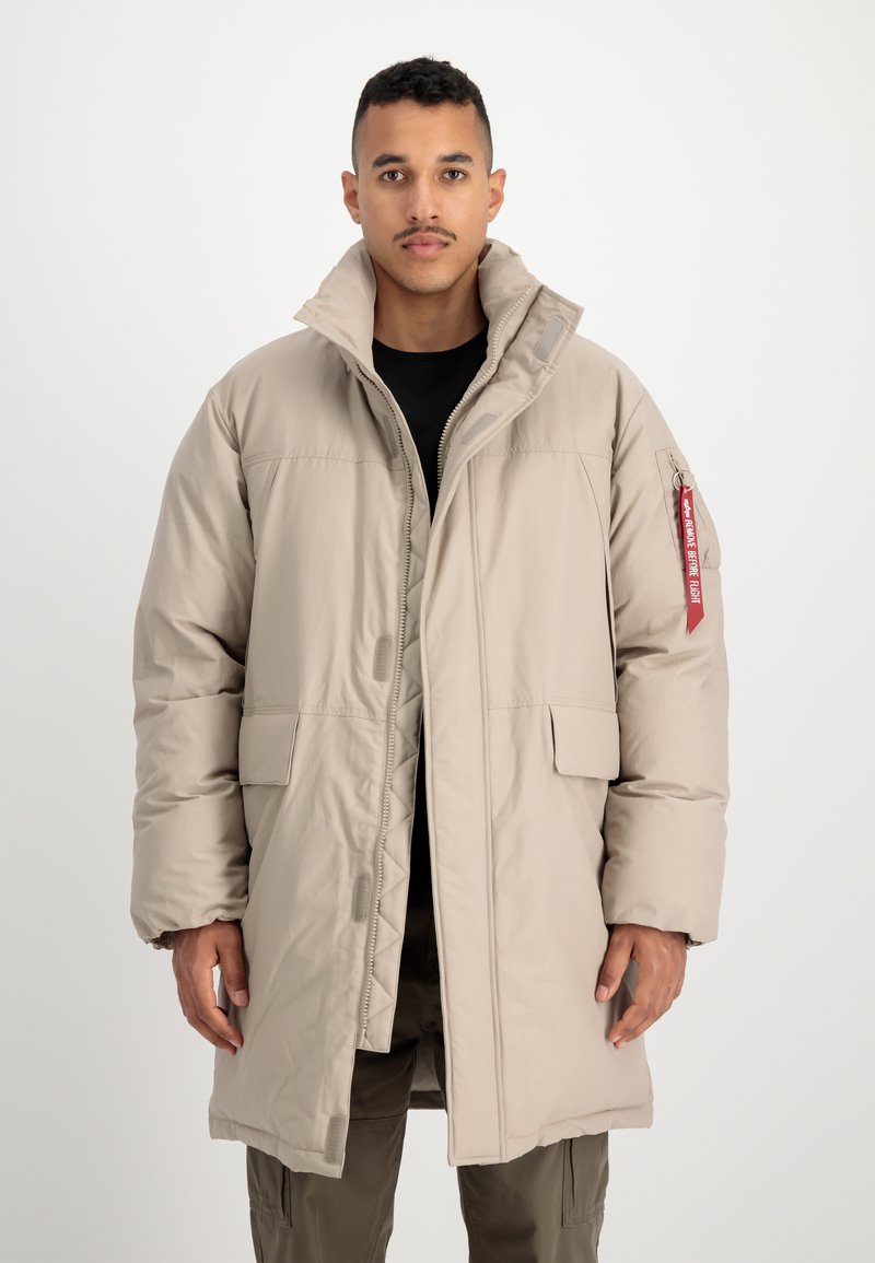 Homme portant un long manteau matelassé beige avec un col haut et une étiquette rouge sur la manche gauche, debout devant un fond blanc uni.