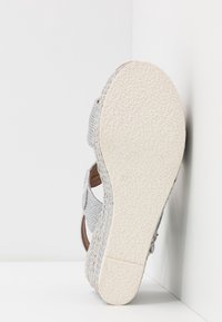 Sandale avec semelle en caoutchouc beige texturée, plateforme compensée tissée et brides en tissu argenté, vue de dessous sur fond blanc.