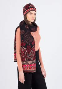 Sorte og røde mønstrede uldsjaal parret med en matchende hue, der har udsmykkede designs og en blød tekstur, båret af en model i en lys top.