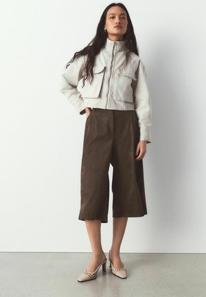 Femeie stând cu mâinile în buzunare, purtând o jachetă crem tăiată scurt, pantaloni largi maro până la jumătatea gambei și pantofi bej cu toc împletiți.