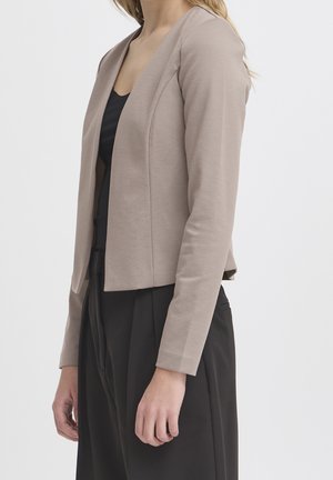 Woman wearing taupe fitted blazer over black top and black high-waisted pants, standing against plain light background.