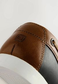 Gros plan du talon d'une basket en cuir marron et noir avec semelle blanche, montrant des coutures détaillées et un logo embossé sur le cuir marron.