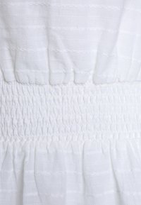 Biała bawełniana sukienka z teksturowanymi paskami i marszczoną talią, z falbanami u dołu. Miękki, lekki materiał.