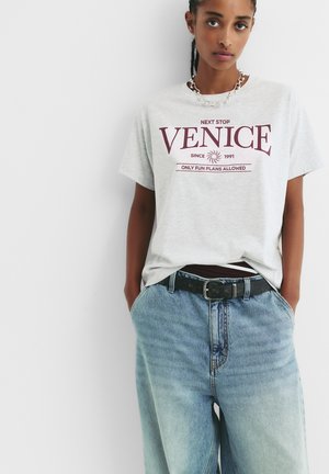T-shirt en coton gris avec une imprimé bordeaux portant les inscriptions "PROCHAIN ARRÊT VENISE" et "SEULES LES PLANS AMUSANTS SONT AUTORISÉS". Associée avec un jean en denim bleu clair et une ceinture noire.