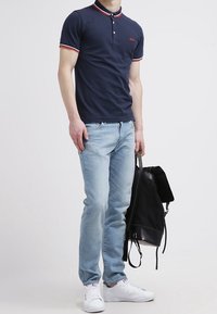 Granatowa koszulka polo z czerwonymi i białymi akcentami, jasnoniebieskie dżinsy, białe sneakersy i czarny plecak. Casualowy strój o dopasowanym kroju.