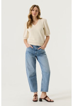 Donna in piedi con le mani nelle tasche, indossa una maglia color crema con scollo a V e maniche a sbuffo, jeans a vita alta azzurri e sandali con tacco marroni.