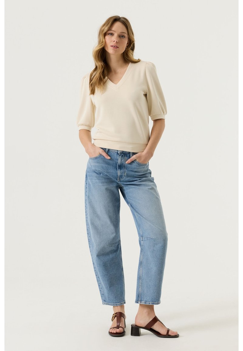 Femme debout, les mains dans les poches, portant un haut crème à manches bouffantes et col en V, un jean taille haute bleu clair, et des sandales à talons marron.