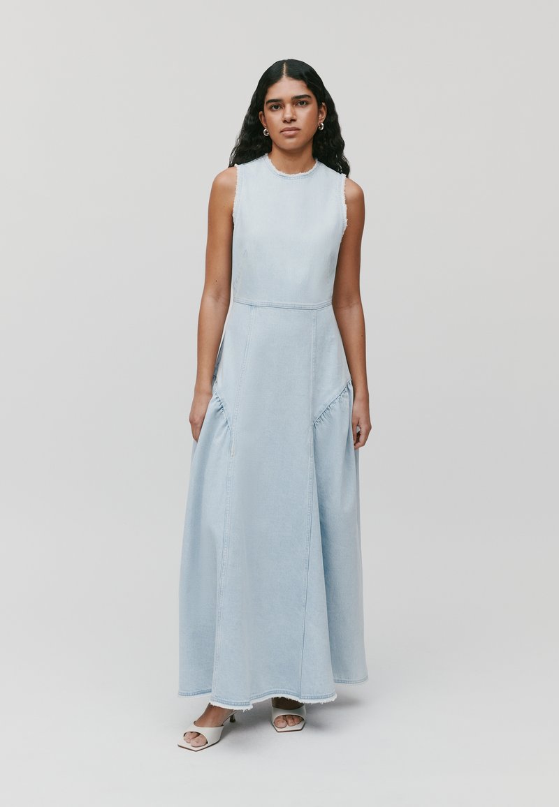 Robe maxi en denim bleu clair avec un col rond, sans manches, détails froncés sur les côtés et ourlet brut. Portée avec des sandales blanches à bout ouvert.