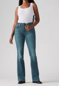 Jeans acampanados azules de mezclilla con una cintura de altura media, ligeras decoloraciones, cinco bolsillos y detalles de costura visibles, combinados con una camiseta sin mangas blanca.