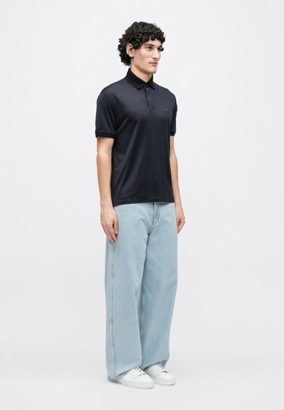 Melns polo krekls ar īsām piedurknēm, apkakli un nelielu logotipu, kopā ar gaiši zilām plašām džinsa biksēm un baltiem sporta apaviem.