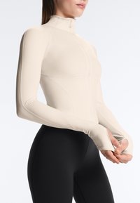 Woman wearing a fitted beige long-sleeve top with thumbholes and high collar, paired with black leggings, against a light background.