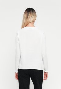 Blonde woman with straight hair wearing a plain white long-sleeve shirt and black pants, standing facing away against a white background.