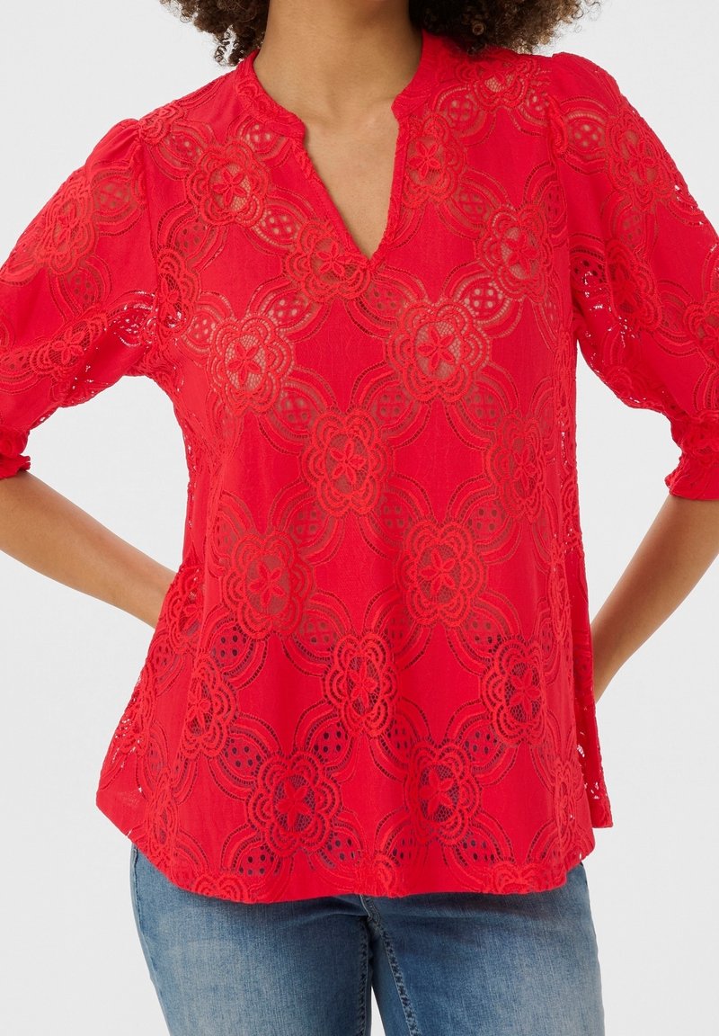 Blusa rossa in pizzo con scollatura a V e maniche a tre quarti, caratterizzata da motivi floreali e dettagli in pizzo aperto in tutto il capo.