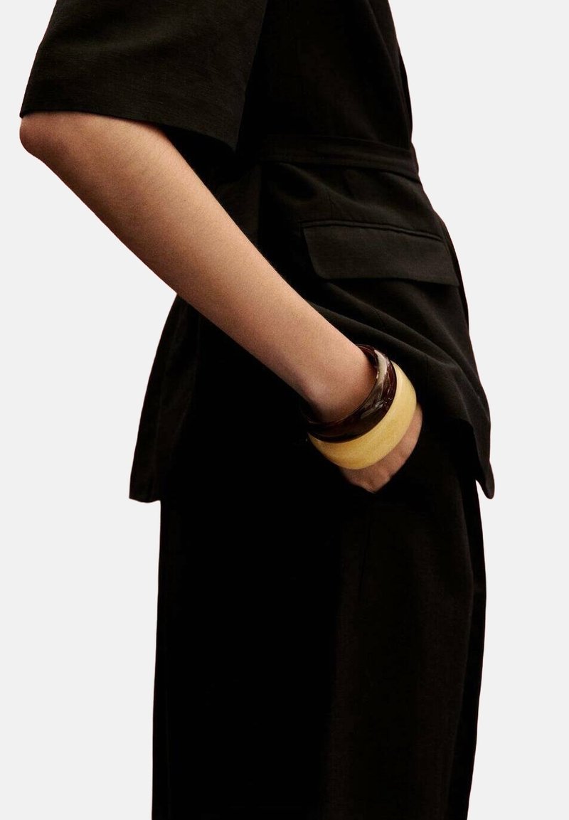 Ręka zgięta, z dłonią w kieszeni, ubrana w czarny strój i dwa szerokie bransoletki, jedna ciemnobrązowa, a druga jasnobeżowa.