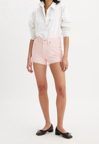 Shorts en denim rose clair avec un ourlet effiloché, présentant des poches avant et une fermeture à bouton. Associés à une chemise blanche à boutons et des ballerines noires.
