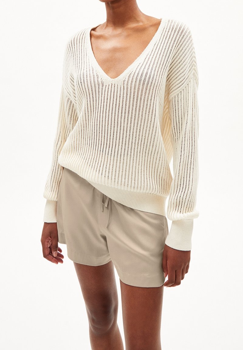Pull en maille crème à col en V avec maille ouverte, manches longues et poignets côtelés, associé à un short beige clair, créant un look décontracté.