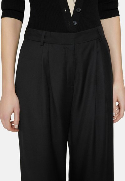 Woman wearing high-waist, loose black trousers and a black long-sleeve top with buttons, hands relaxed by sides.