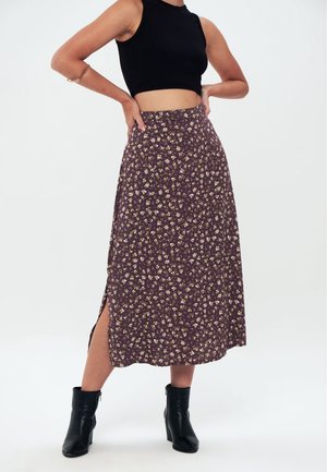 Vrouw draagt een zwart mouwloos crop-top, paarse midi-rok met bloemenprint en een zijsplit, zwarte enkellaarsjes en een gouden armband aan haar linkerarm.