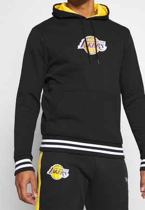 Person trägt schwarzen Los Angeles Lakers Hoodie und passende Hose mit gelben Akzenten und Teamlogos auf Brust und Oberschenkel.