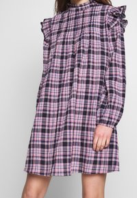 Vestido a cuadros con hombros con volantes, cuello alto, mangas largas y una silueta acampanada en un patrón de cuadros en tonos rosa y azul marino.