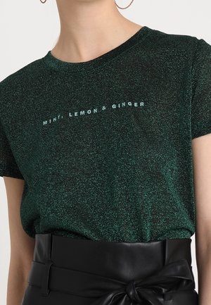 Kobieta w błyszczącej ciemnozielonej koszulce z napisem "MINT, LEMON & GINGER", włożonej w wysokowalne czarne skórzane spodnie z paskiem wiązanym w kokardę.