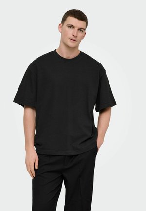 Ung mand med kort hår iført en løs sort T-shirt og sorte bukser, stående med den ene hånd i lommen mod en hvid baggrund.