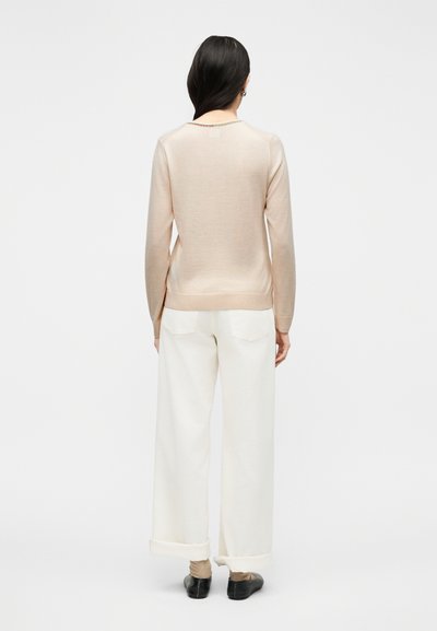 Γυναίκα με μακριά σκούρα μαλλιά, ντυμένη με μπεζ πουλόβερ, λευκό παντελόνι με φαρδιά μπατζάκια και μανσέτες, καφέ κάλτσες και μαύρα παπούτσια χωρίς κορδόνια, όρθια με την πλάτη προς τα εμάς.