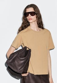 Beige Baumwoll-T-Shirt mit kurzen Ärmeln, kombiniert mit einer großen dunkelbraunen Lederhandtasche. Model trägt übergroße schwarze Sonnenbrille.