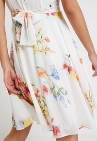 Vestito floreale in tessuto leggero e trasparente con una base bianca. Presenta una cintura con nodo e una stampa colorata di vari fiori in rosso, giallo e blu.